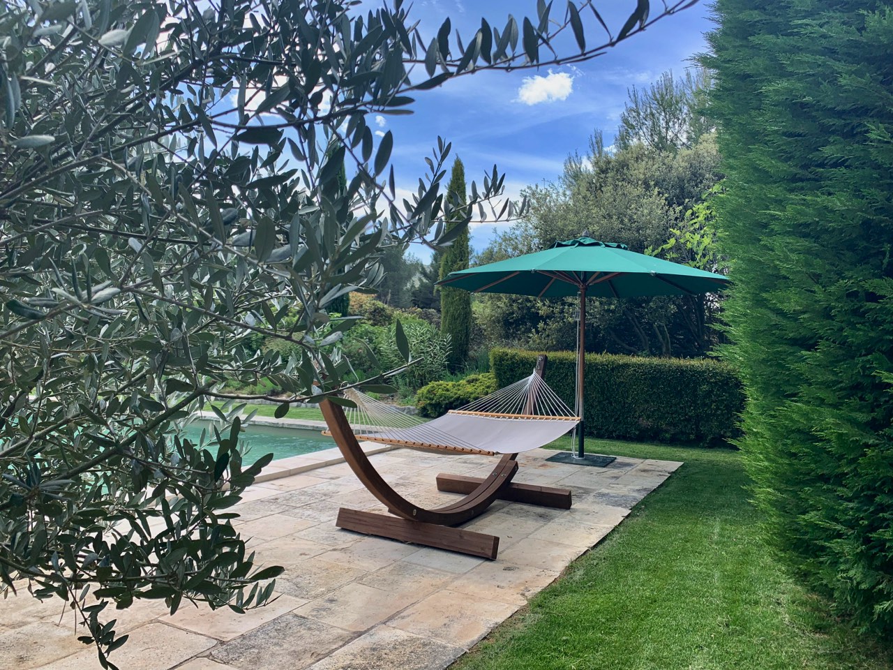 A tranquil garden scene featuring a hammock under a green umbrella on a stone patio, surrounded by lush greenery and trees, with a glimpse of a swimming pool on the left.